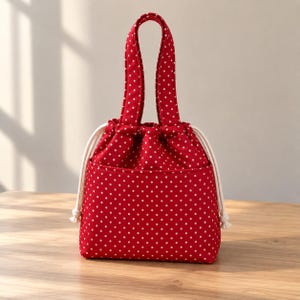 May include: A red drawstring bag featuring a white polka dot pattern throughout. The bag includes two matching fabric handles and thick, cream-coloured rope drawstrings on both sides that cinch the top opening closed for secure storage.