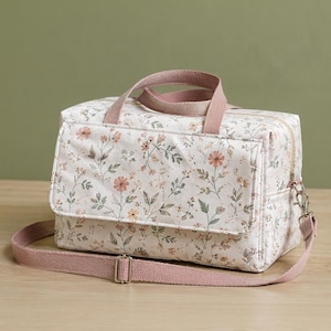 May include: A rectangular fabric bag featuring a white base with a delicate floral pattern in shades of pink, orange, and green. The bag includes a top handle, a front flap closure, and an adjustable pink shoulder strap with metal hardware.