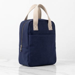 May include: A navy blue rectangular lunch bag made of canvas material. It features two beige woven fabric handles attached to the top, a zippered main compartment closure, and a flat front pocket. The bag has a structured, upright design.