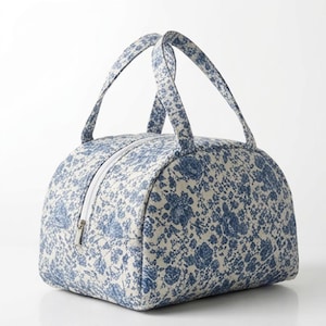 May include: A dome-shaped lunch bag featuring a blue and white floral pattern. The bag has two fabric handles on top and a white zipper closure that runs along the center. The exterior is made of a quilted fabric with a repeating botanical print.