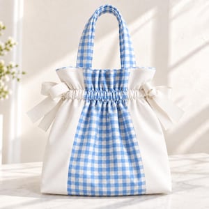 Puede incluir: Un bolso tote con un patrón de cuadros vichy en azul y blanco en el panel central y las asas, contrastado por paneles laterales en blanco sólido. El bolso incluye un cierre superior fruncido con lazos de cinta blanca en cada lado.