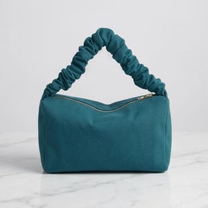 May include: A teal canvas handbag featuring a rectangular body and a single gathered, scrunchie-style handle. The bag includes a top zipper closure with a metallic pull, showcasing a solid color design with a textured fabric finish throughout.