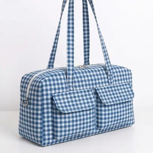 Puede incluir: Un bolso de viaje rectangular con un patrón de cuadros vichy en azul y blanco. El bolso incluye dos bolsillos exteriores frontales con solapas, un cierre de cremallera blanco en la parte superior y dos correas largas para el hombro.