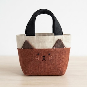 May include: A small tote bag featuring a two-tone design with a rust-colored lower section and a cream-colored upper section. The bag includes two black fabric handles, two dark brown triangular ears, and embroidered black cat facial features.