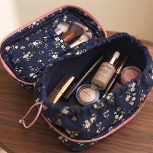 May include: A navy blue fabric cosmetic bag with a white and yellow floral pattern, featuring a pink zipper and drawstring closure. The open bag contains various makeup items including brushes, a foundation bottle, and small round compacts.