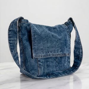 May include: A medium blue acid-wash denim shoulder bag featuring a rectangular front flap closure, a wide integrated shoulder strap, and visible topstitching details throughout the structured fabric construction of the bag.