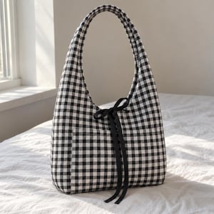 May include: A hobo-style shoulder bag featuring a black and white gingham check pattern. The bag has a single integrated handle, a front exterior pocket, and a decorative black ribbon tied into a bow at the centre of the front panel.