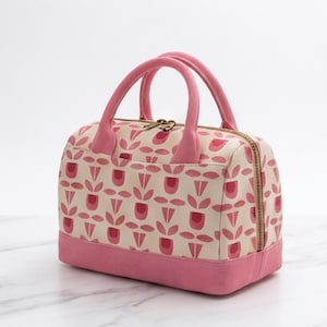 Könnte beinhalten: Eine rechteckige Lunchtasche mit zwei rosa Griffen und einem Reißverschluss. Die Tasche hat einen cremefarbenen Stoffkörper mit einem sich wiederholenden Muster aus stilisierten rosa tulpenartigen Blumen und einem festen rosa Boden.