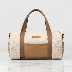 Op de afbeelding: Een cilindervormige plunjezak met een combinatie van crèmekleurige sherpa-fleece en beige kunstlederen panelen. De tas is voorzien van twee beige webbing handvatten, een centraal buitenvak en een klein rechthoekig merklabel aan de voorzijde.