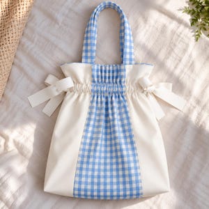 May include: A tote bag featuring a central vertical panel of blue and white gingham fabric, flanked by solid off-white side panels. The bag includes matching gingham handles, a gathered drawstring-style top, and decorative off-white ribbon bows.