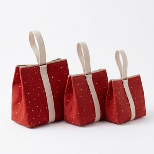 Puede incluir: Tres bolsas de lona roja de diferentes tamaños, cada una con un patrón de puntos blancos y una sola asa de tela beige unida a una tira vertical beige en el centro del panel frontal. Las bolsas tienen una base rectangular estructurada.