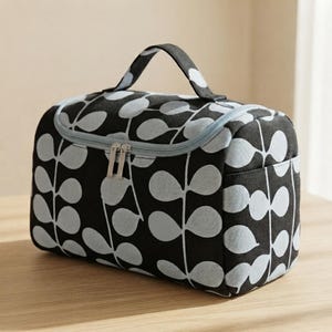 May include: A rectangular toiletry bag featuring a black fabric base with a repeating light grey leaf pattern. The bag includes a top handle, a zippered main compartment with two silver-toned metal zipper pulls, and a light grey fabric trim.