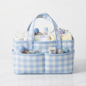 May include: A rectangular fabric storage caddy featuring a light blue and white gingham check pattern. The caddy has two sturdy handles and two exterior front pockets. It is filled with various baby care items, including white cloths, a small bottle, and toys.