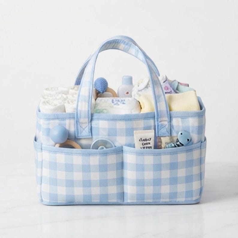 May include: A rectangular fabric storage caddy featuring a light blue and white gingham check pattern. The caddy has two sturdy handles and two exterior front pockets. It is filled with various baby care items, including white cloths, a small bottle, and toys.