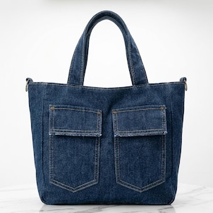 May include: A dark blue denim tote bag featuring two front pockets with frayed edges, metallic rivets, and two top handles. The bag has a structured rectangular shape with visible stitching and side metal rings for an optional shoulder strap.