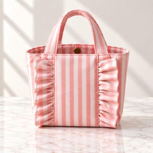 Può includere: Una piccola borsa tote con un motivo a righe verticali rosa e bianche. La borsa presenta due manici superiori e un bordo in tessuto arricciato che corre verticalmente sul pannello frontale. Un bottone a pressione in metallo dorato è visibile.