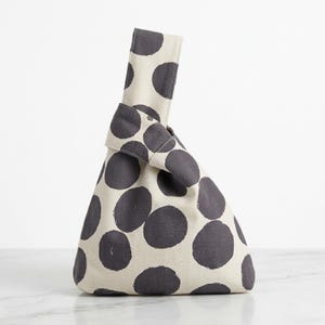 May include: A Japanese-style knot bag made of off-white canvas fabric featuring a pattern of large, dark gray, hand-painted style polka dots. The bag has a unique asymmetrical handle design where one strap pulls through the other to close.