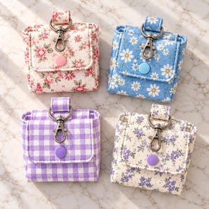Puede incluir: Cuatro bolsos cuadrados pequeños con cierres de mosquetón metálico y botones a presión. Presentan estampados variados: flores rosas, margaritas sobre fondo azul, cuadros vichy morados y flores pequeñas moradas sobre fondo crema.