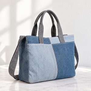 Op de afbeelding: Een rechthoekige draagtas gemaakt van patchwork denimstof in verschillende blauwtinten. De tas is voorzien van twee handvatten van donkergrijze denim en een bijpassende schouderriem van donkergrijze denim die aan de zijkanten is bevestigd.