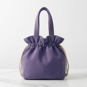 Könnte beinhalten: Eine violette Kordelzugtasche aus Cord mit gerafftem Oberrand, zwei passenden Stoffhenkeln und dicken beigen Kordelzügen aus Seil an beiden Seiten. Die Tasche weist eine vertikale Rippenstruktur auf der gesamten Außenfläche auf.