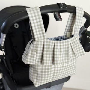 Könnte beinhalten: Eine Kinderwagen-Organizer-Tasche aus hellgrau-weißem Vichy-Stoff mit einer gerüschten Vorderseite und zwei Tragegriffen. Die Tasche ist am schwarzen Lenker eines Kinderwagens befestigt und zeigt ein Innenfutter mit dunkelblau-weißem Karomuster.
