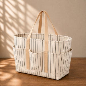 May include: A rectangular canvas tote bag featuring a cream-colored base with thin vertical black stripes. The bag includes two light beige fabric handles and three exterior pockets on the front, providing organized storage space for various items.