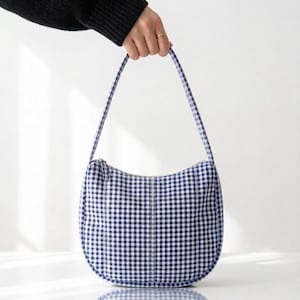 May include: A crescent-shaped shoulder bag featuring a blue and white gingham check pattern. The bag has a single integrated strap, a curved silhouette, and a top zipper closure. The fabric displays a consistent grid design throughout the exterior.