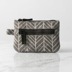 May include: A small rectangular zippered pouch featuring a grey and beige herringbone pattern. The accessory includes a black zipper closure along the top and a smaller horizontal black zippered pocket on the front, complete with a black fabric pull tab.
