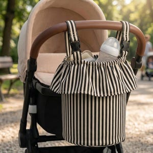 Può includere: Una borsa organizer cilindrica per passeggino con motivo a righe verticali bianche e nere e bordo superiore arricciato. La borsa include due cinghie nere regolabili con fibbie in plastica per il fissaggio al passeggino e un biberon.