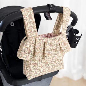 Peut inclure: Un sac organisateur de poussette en tissu beige avec un petit imprimé floral dans des tons rose et vert. Le sac possède deux anses, un panneau avant à volants et une doublure intérieure à carreaux roses et blancs, suspendu à une poignée noire.