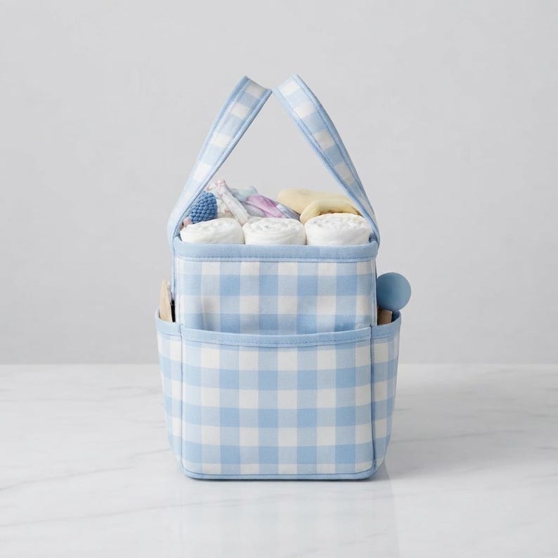May include: A light blue and white gingham fabric storage caddy with two handles and exterior pockets. The interior is filled with several white rolled cloths and small items, including a blue sphere and a wooden object visible on the side.