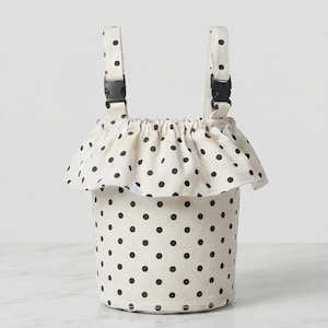May include: A cylindrical fabric bag featuring a cream base with a black polka dot pattern. The bag includes a ruffled top edge and two vertical fabric straps, each equipped with a black plastic side-release buckle for secure attachment.