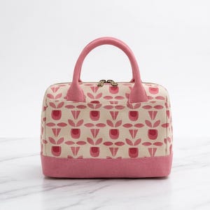Pode incluir: Uma mala retangular com base em tom creme e um padrão repetitivo de formas estilizadas de tulipas em rosa e rosa escuro. A mala inclui duas pegas superiores cor-de-rosa, um painel de base rosa e um fecho de correr com detalhes dourados.