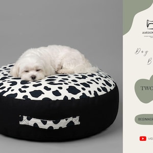 May include: A round dog and cat bed with a black and white cow print top and black sides. A small white dog is sleeping on the bed. The bed has a handle and the text "TWO SIZE" and "BEGINNER FRIENDLY".