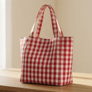 Peut inclure: Un sac cabas avec un motif à carreaux vichy rouge et blanc. Le sac possède deux anses verticales en tissu fixées à l'ouverture supérieure, une base rectangulaire et un corps structuré avec des coutures visibles le long des côtés.