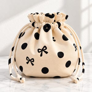 Puede incluir: Una bolsa de tela color crema con cierre de cordón, decorada con un patrón repetitivo de lunares negros y pequeños lazos negros. La bolsa tiene una parte superior fruncida asegurada por dos cordones trenzados gruesos de color crema.