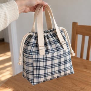 May include: A cream-colored fabric handbag featuring a dark blue and black plaid pattern. The bag includes two sturdy cream-colored canvas handles and a drawstring closure with thick cream-colored rope cords on both sides for secure storage.