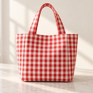 May include: A tote bag featuring a vibrant red and white gingham check pattern. The bag has two matching fabric handles attached to the top, a rectangular base, and a structured body with clean vertical seams along the front and sides.