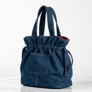 Può includere: Una borsa a mano in denim blu scuro con chiusura a coulisse arricciata, due robusti manici e una tasca esterna rettangolare sul davanti. La fodera interna è di un colore magenta intenso, visibile nell'apertura superiore.