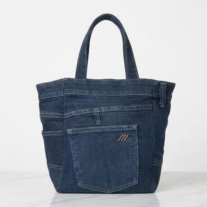 May include: A dark blue denim tote bag featuring two top handles, a large exterior patch pocket on the front, and a small metallic logo emblem with three diagonal lines attached to the pocket. The bag has visible stitching and seams.
