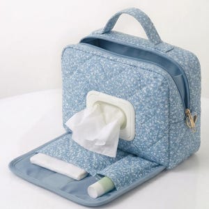 Puede incluir: Un estuche de viaje acolchado de color azul claro con estampado floral blanco, que cuenta con un asa superior, cierre de cremallera y una abertura central de plástico con una toallita blanca. El interior incluye un bolsillo plano y un objeto cilíndrico.