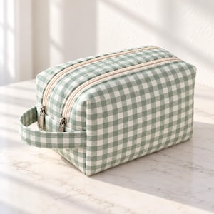 Könnte beinhalten: Eine rechteckige Kulturtasche mit einem salbeigrünen und weißen Vichy-Karo-Muster. Die Tasche verfügt über zwei Reißverschlüsse an der Oberseite mit silberfarbenen Metallschiebern und einen passenden Stoffgriff an der Seite zum Tragen.