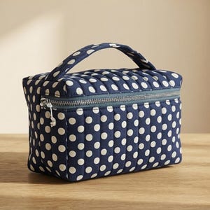 Puede incluir: Un neceser rectangular acolchado de color azul marino con un estampado de lunares blancos. El bolso incluye un asa superior, un cierre de cremallera horizontal con un tirador de metal plateado y un diseño estructurado con costuras verticales.
