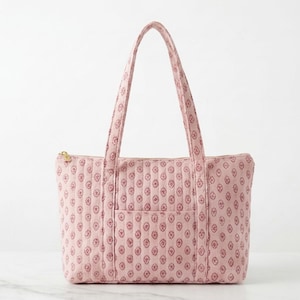 May include: A quilted tote bag in a soft pink hue featuring a repeating dark pink diamond-shaped floral pattern. The bag includes two long shoulder straps, a top zip closure with a gold-toned metal pull, and a single exterior slip pocket.