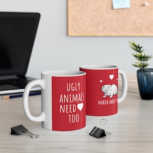 May include: Two white ceramic mugs with red panels. One mug has the text "UGLY ANIMALS NEED TOO" and a heart. The other mug has a cartoon naked mole-rat and the text "NAKED MOLE-RAT".