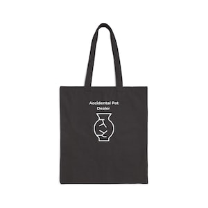 May include: A black tote bag with white text that reads "Accidental Pot Dealer" above a white line drawing of a vase. The bag has a rectangular shape with a handle attached at the top.