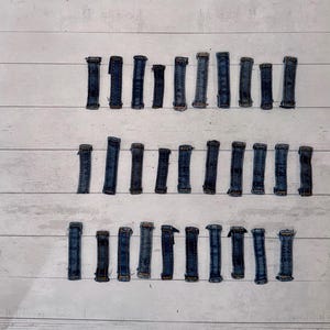 May include: A collection of denim jean belt loops arranged in three rows on a white wooden surface. The loops are various lengths and shades of blue, showing signs of wear.