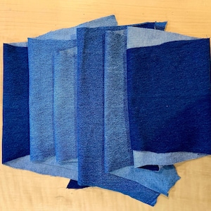 May include: Assortment of denim fabric pieces in various shades of blue, arranged in a layered pattern. The fabric appears to be new, with a range of indigo hues from light to dark.