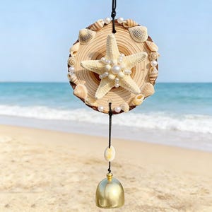 May include: A decorative wind chime featuring a wooden disc adorned with seashells and a starfish embellished with pearls. A gold-colored bell hangs below, suspended by a black cord. The background shows a beach and ocean.