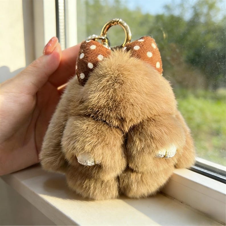 May include: A fluffy, light brown bunny keyring with a brown polka dot bow and a gold-coloured metal ring. The keyring has rhinestone accents on the feet.